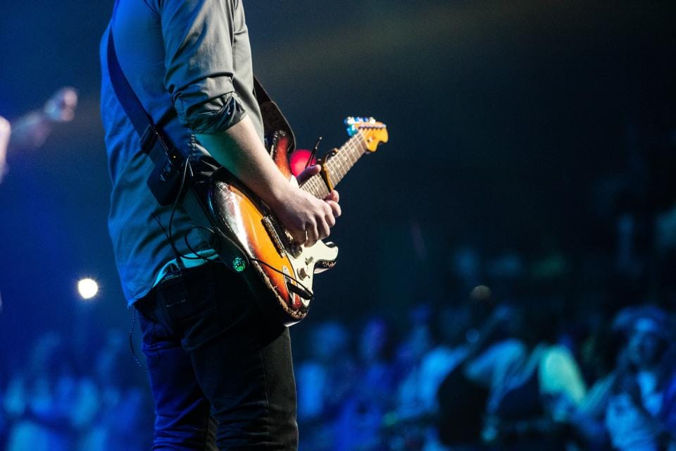 Guitarrista visto de costas tocando em um palco, com a plateia iluminada de azul ao fundo
